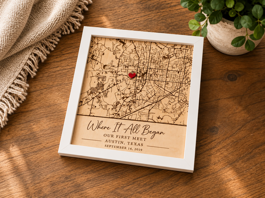 A square framed wooden map print with a white frame sits on a wooden table. The map is engraved with fine lines and a small red heart near the center, with the text 'Where It All Began Our First Meet Austin, Texas September 14, 2015' at the bottom.
