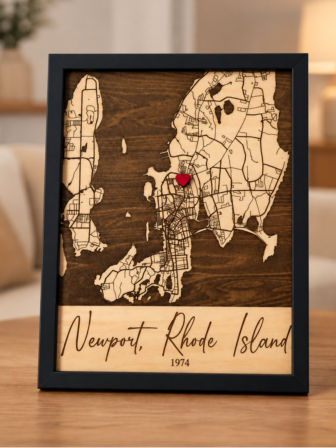A framed wooden map of Newport, Rhode Island, featuring a dark brown wood background, light beige map outlines, and a small red heart pin near the center, displayed on a wooden table in a softly lit living room.