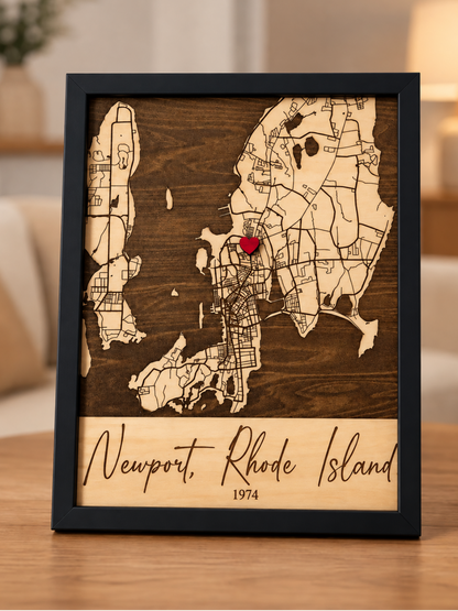 A framed wooden map of Newport, Rhode Island, featuring a dark brown wood background, light beige map outlines, and a small red heart pin near the center, displayed on a wooden table in a softly lit living room.